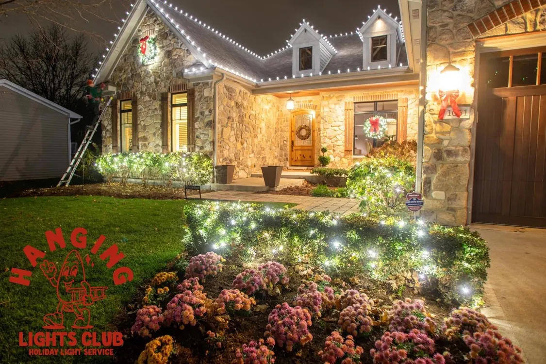 Residential house with white holiday lights on the roofline and bushes, with a person on a ladder installing lights.