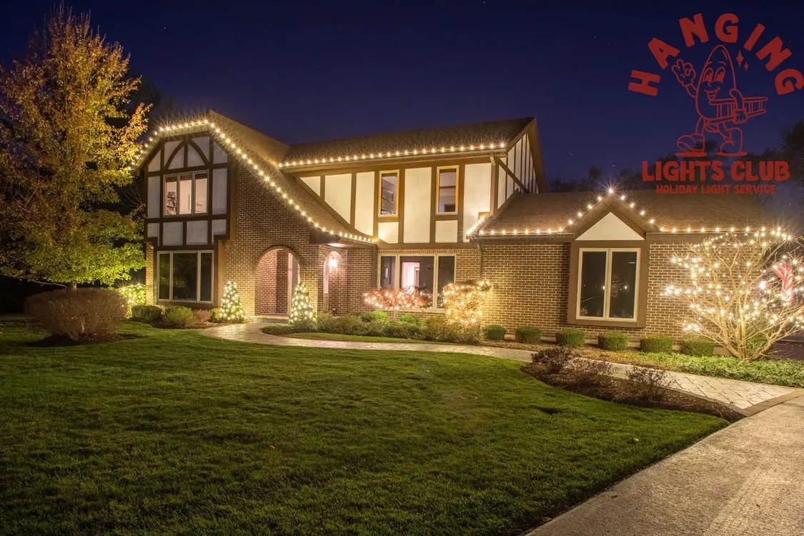 Large brick house with warm white holiday lights on the roofline and small trees, in Tower Lakes
