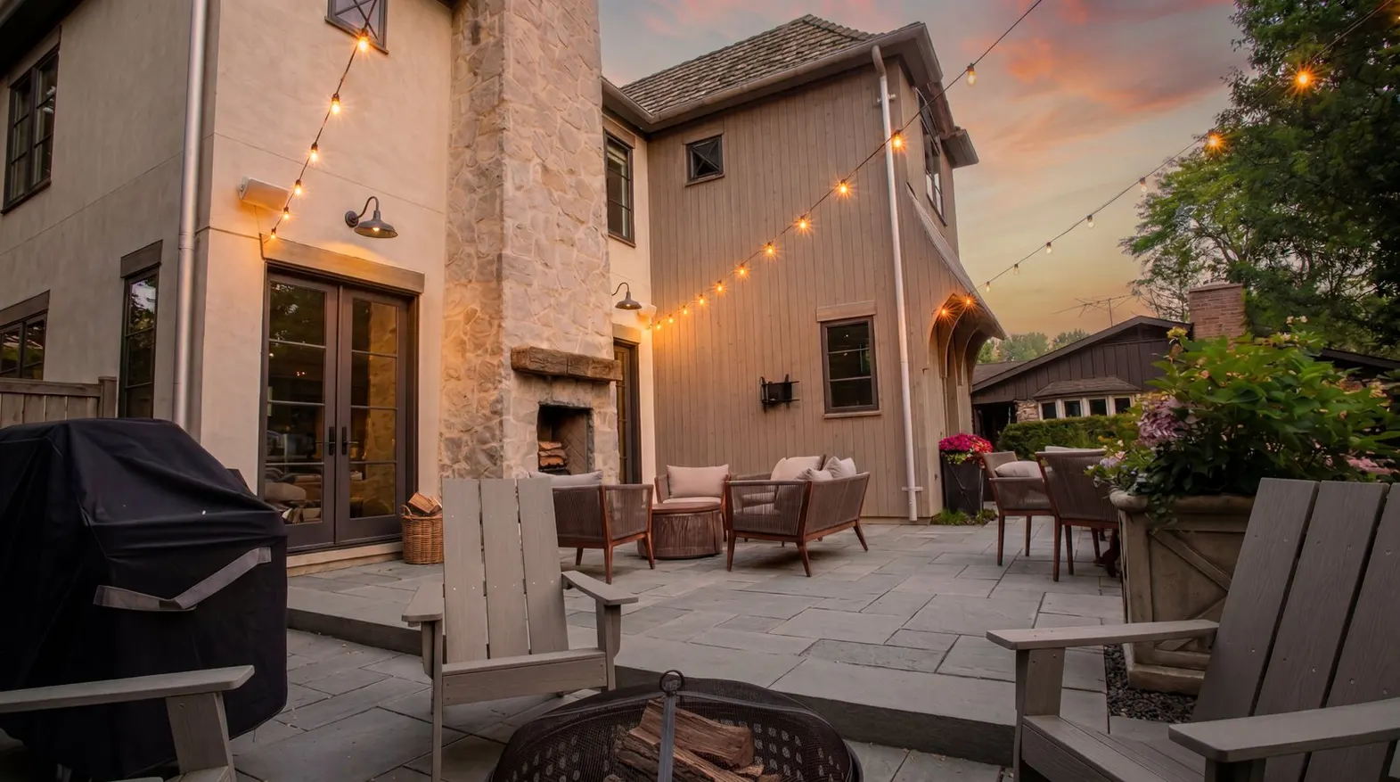 Beautiful patio at sunset with warm string lights overhead, outdoor seating, fireplace, and lush landscaping