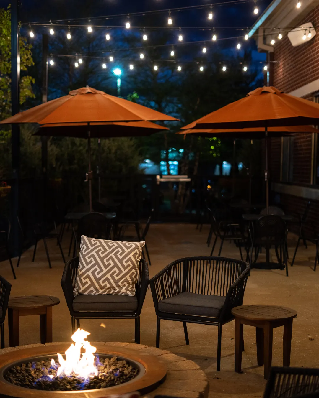 Elegant commercial patio at night with string lights overhead, orange umbrellas, fire pit in foreground, comfortable seating with patterned pillows, and ambient blue accent lighting in the background
