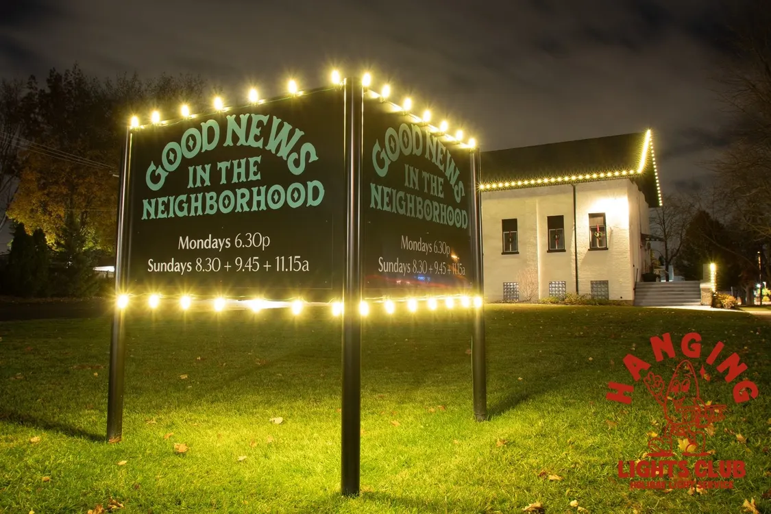 House with warm white lights on the roofline and a 'GOOD NEWS IN THE NEIGHBORHOOD' sign