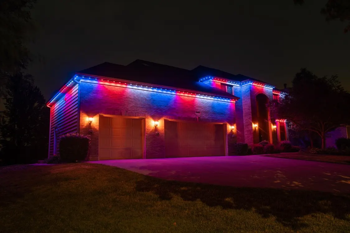 House with vibrant multicolor permanent lighting - blue and red LED lights illuminating the roofline, garage doors, and architectural features at night